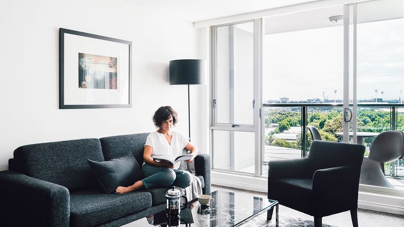 Lady reading a magazine in 1 Bedroom Deluxe Apartment lounge at Quay West Suites Melbourne