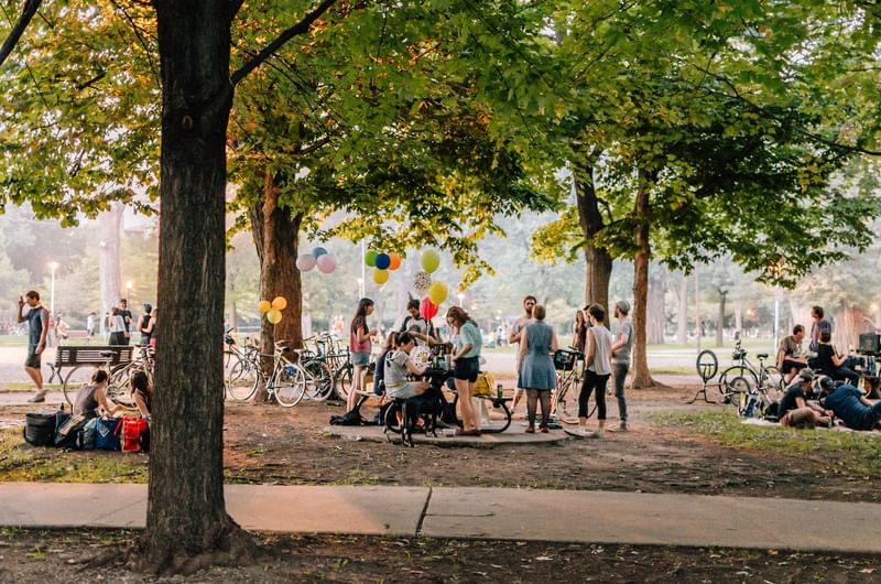 People gather with colorful balloons and bicycles under large trees in a lively park at Warwick Le Crystal Montreal