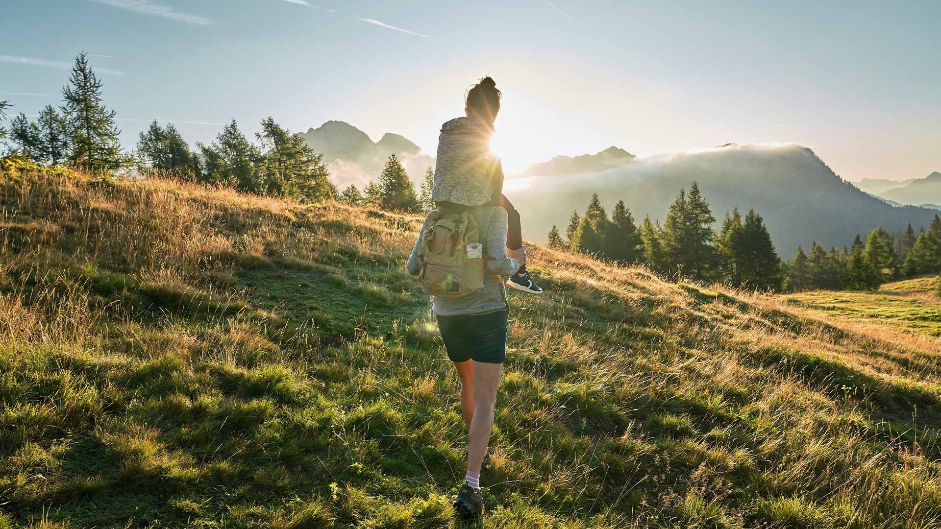 Woman hiking with a child on her back in a meadow with mountains in the background.