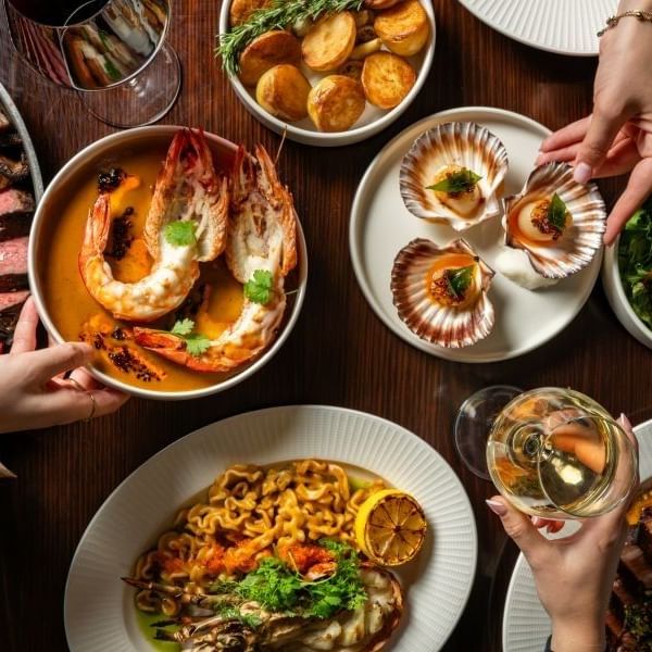 Overhead view of a shared dining table featuring sliced steak, grilled prawns, seared scallops, roasted potatoes, and greens.