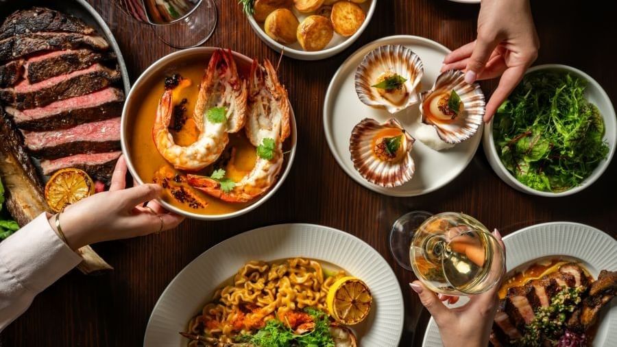 Overhead view of a shared dining table featuring sliced steak, grilled prawns, seared scallops, roasted potatoes, and greens.