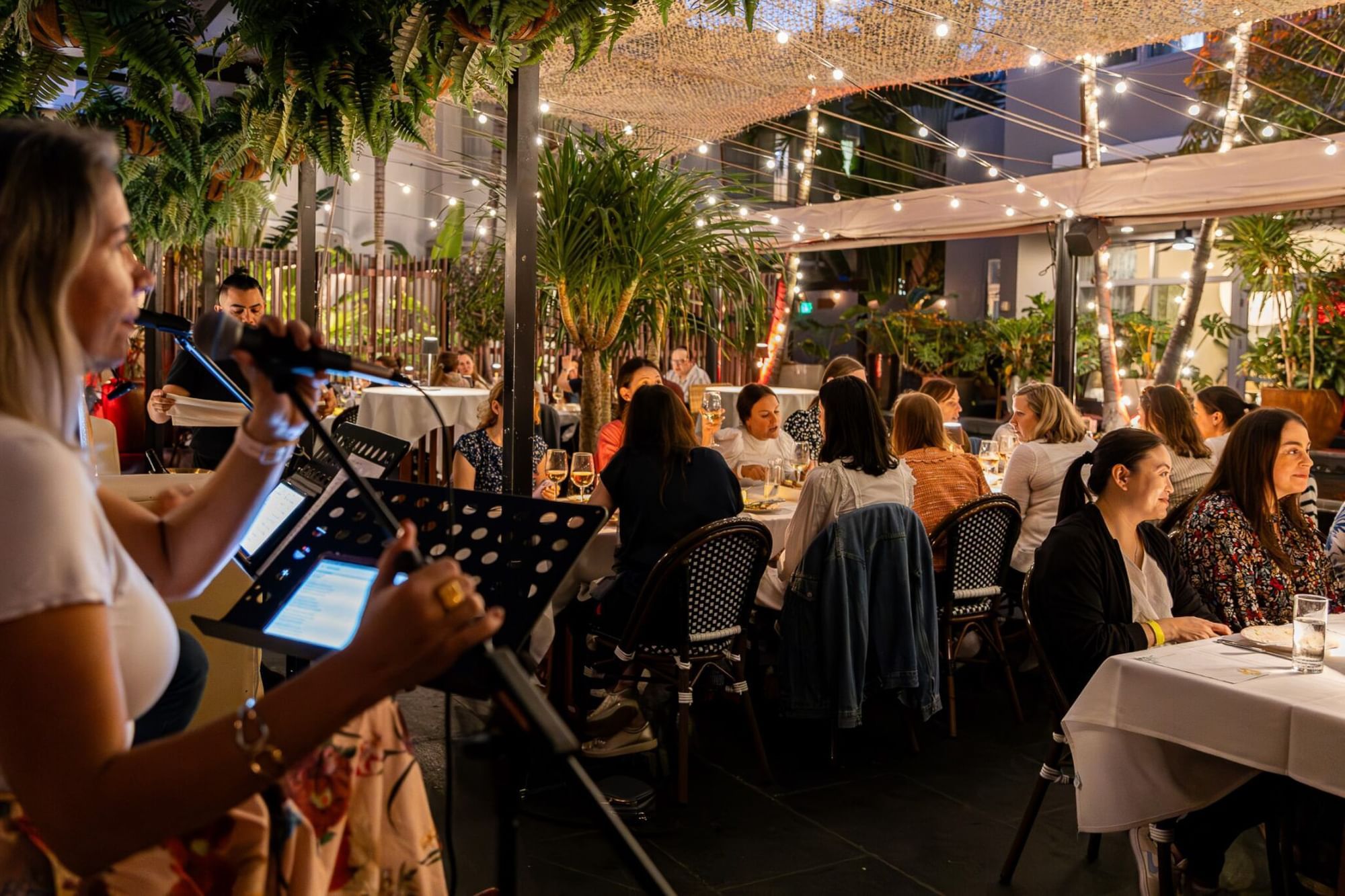 a woman sings into a microphone at a dining area with people sitting at tables