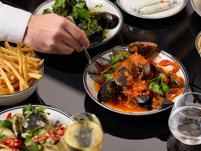 Hand serving mussels and fries with sauces and garnishes in bowls and glasses.