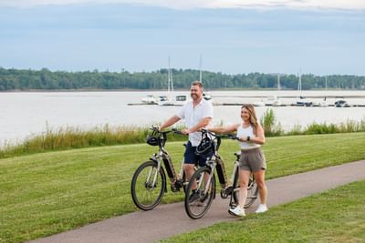Couple walking with bicycles on a paved path by a grassy field near a calm bay at Rodd Brudenell River Resort