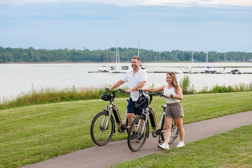 Couple walking with bicycles on a paved path by a grassy field near a calm bay at Rodd Brudenell River Resort