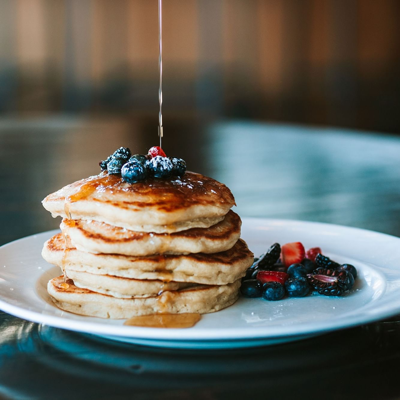 Stack of pancakes with berries and maple syrup being poured on top.