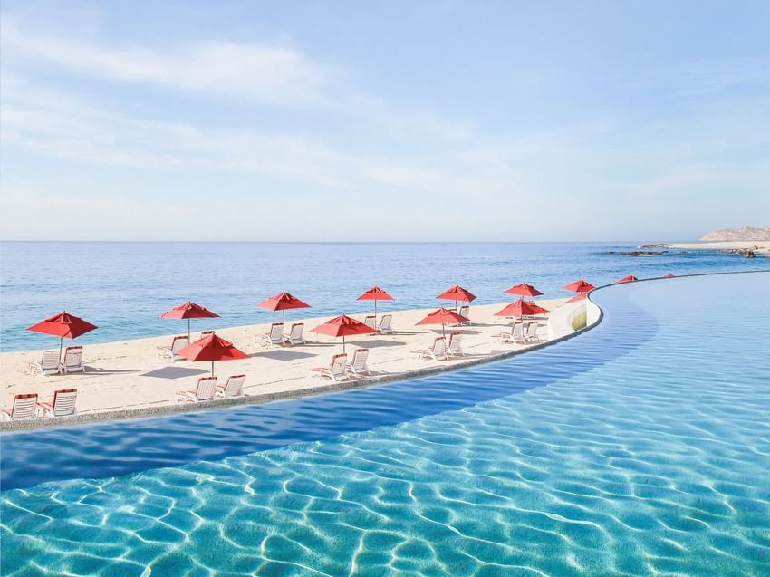 A stunning infinity pool merges with the ocean at Marquis Los Cabos Resort with red umbrellas lining the sandy edge