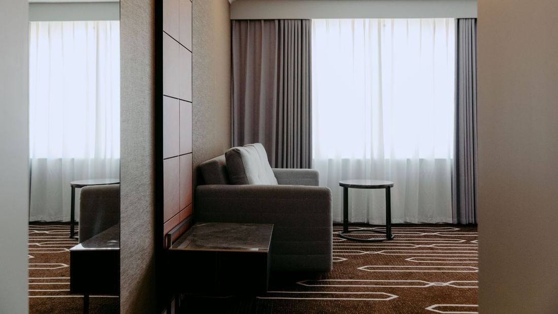 Hotel room with gray couch, two pillows, side tables, and large window with curtains.