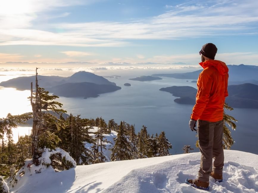 A man in winter gear enjoys a snowy mountain view with lake and hills below for Prepaid Mastercard Promotion.