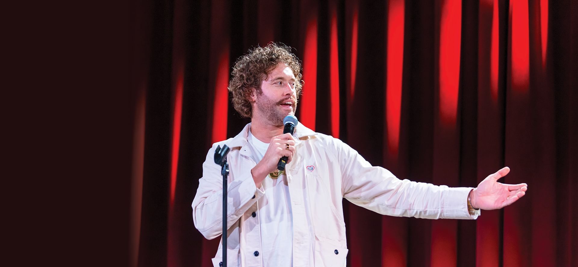 Actor and comedian T.J. Miller with microphone gesturing on stage in front of red curtain backdrop at The Brook
