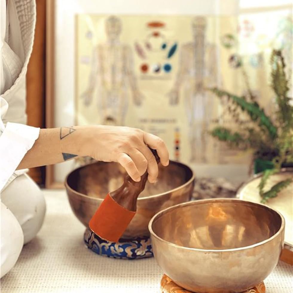 Person mit tätowiertem Handgelenk spielt auf zwei tibetischen Klangschalen während des Five Elements Sound Rituals.