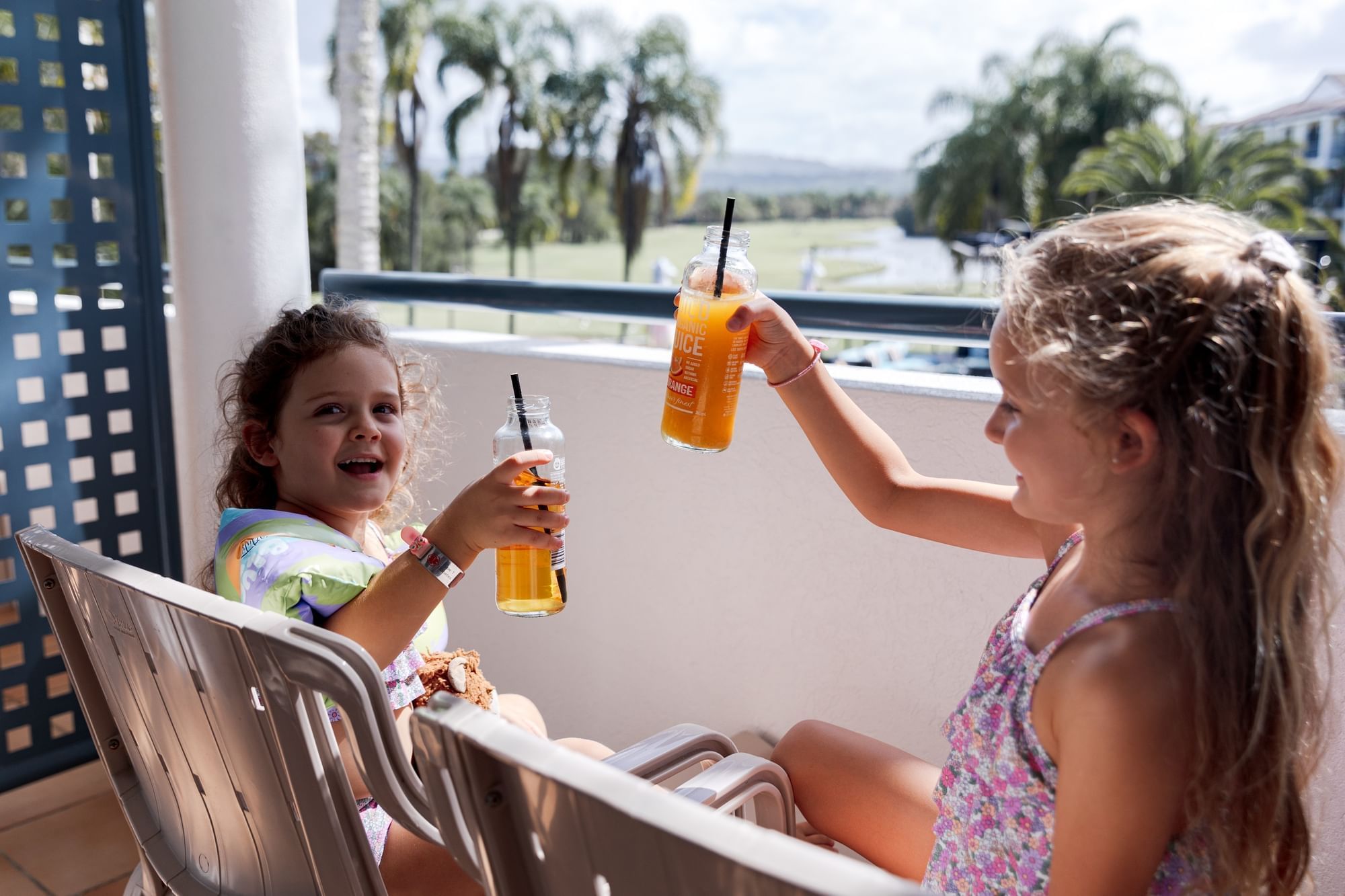 Two girls on a balcony with a view of trees and buildings, holding juice bottles and laughing.
