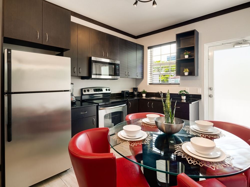 A kitchen with a dining area, featuring a glass table with red chairs, stainless steel appliances, and dark cabinets.