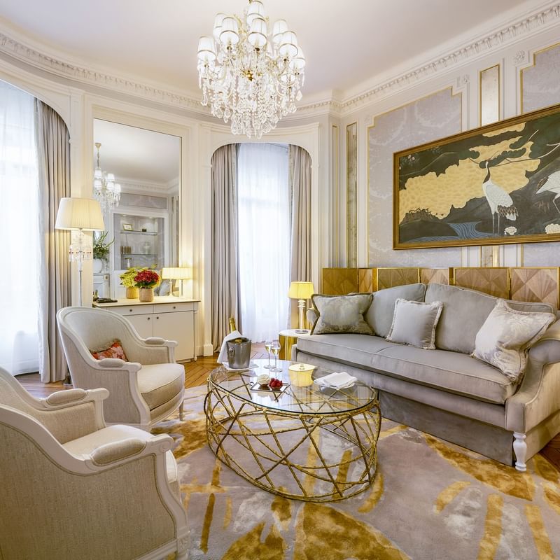 Elegant living room with gray couch, chairs, glass table, chandelier, artwork, and lamps.