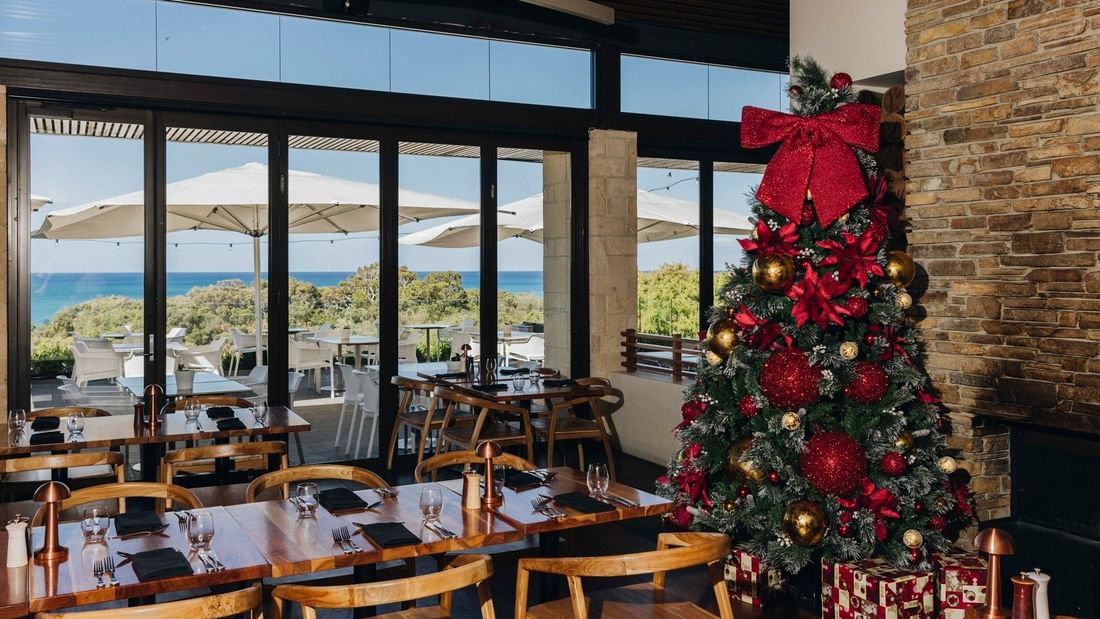 Restaurant dining area with decorated Christmas tree, ocean views through large windows, and festive table settings