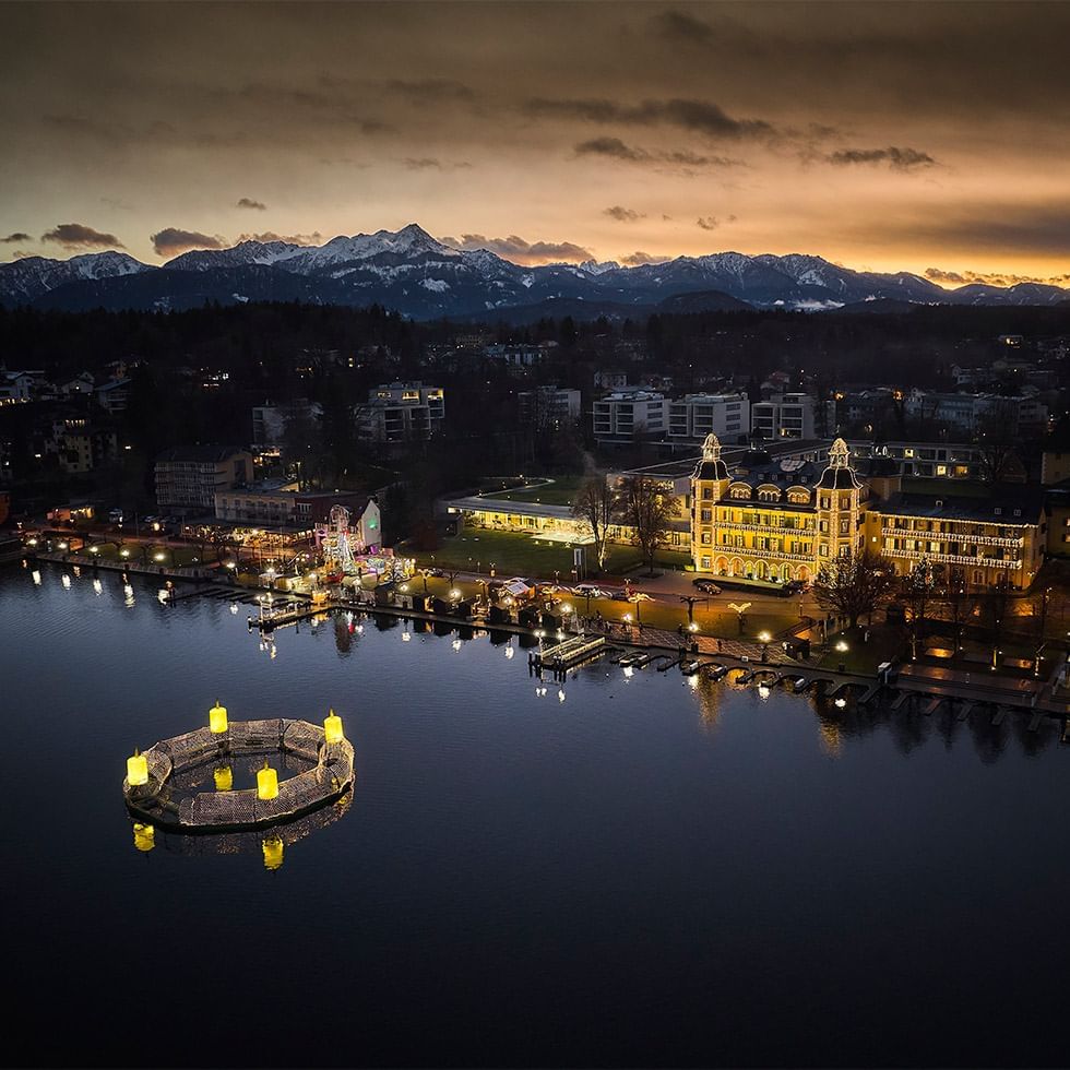 Vista aerea al tramonto di una città con edifici illuminati, monti innevati sullo sfondo e un lago.