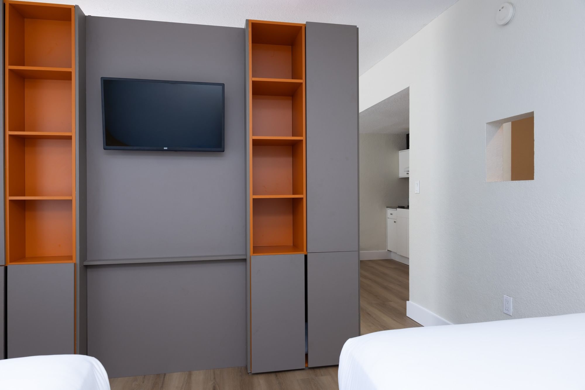 A studio room with a wall-mounted TV, shelves, and a kitchenette in the background.