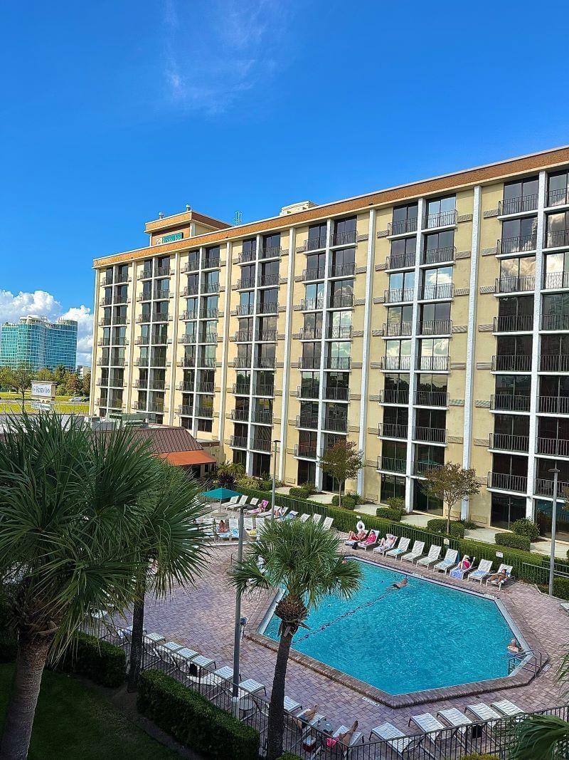 The exterior of Rosen Inn Closest to Universal hotel showing dozens of room windows, the pool, white chairs on the pool deck, and palm trees. 