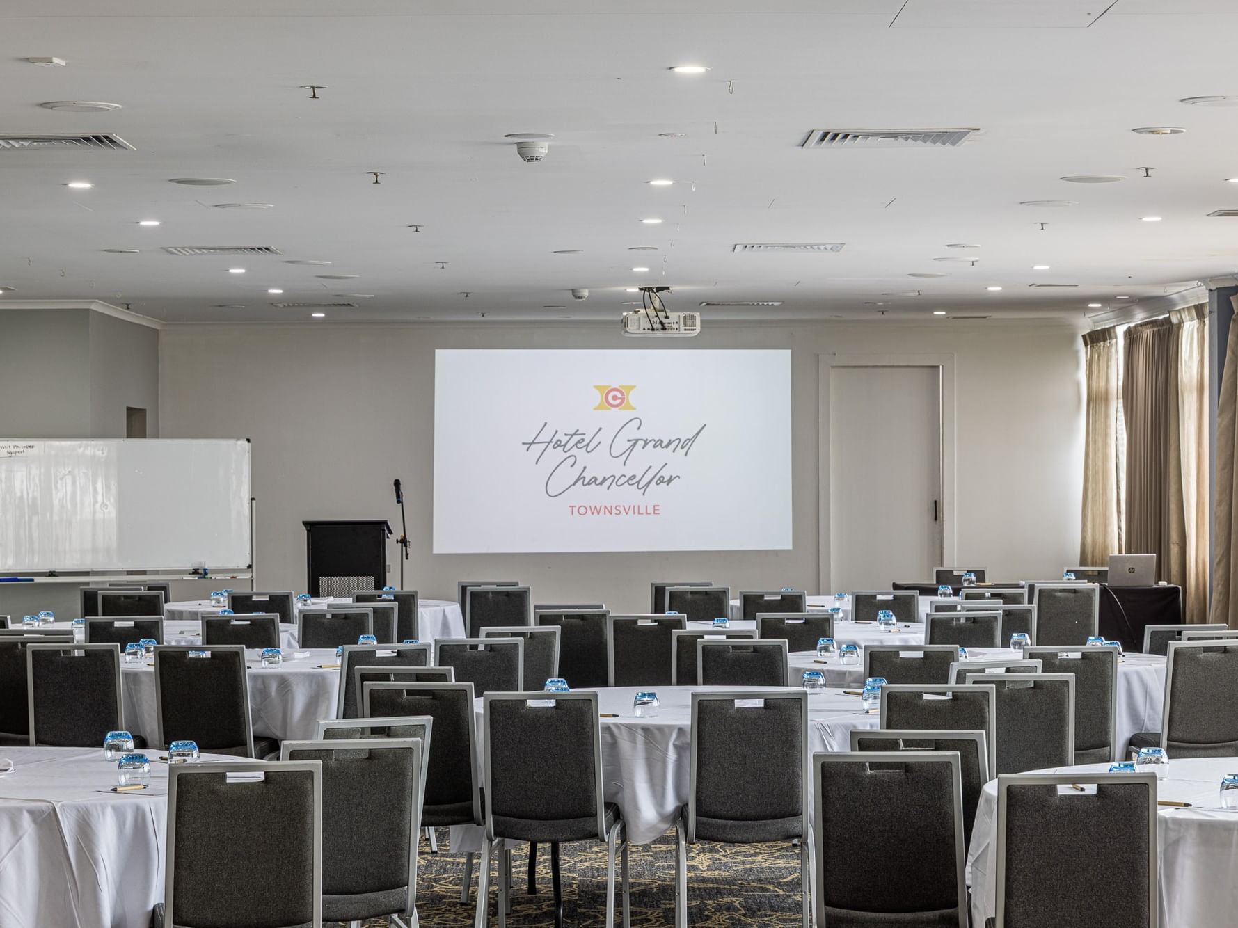 A spacious conference room set up with round tables, grey chairs, a whiteboard, and a large projector screen displaying the hotel logo.