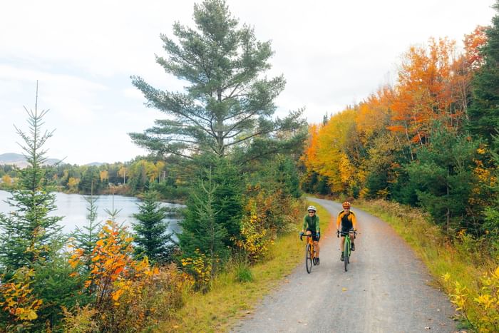Two cyclists ride on a scenic dirt path beside a lake surrounded by autumn trees near Lake Placid Club Grandview