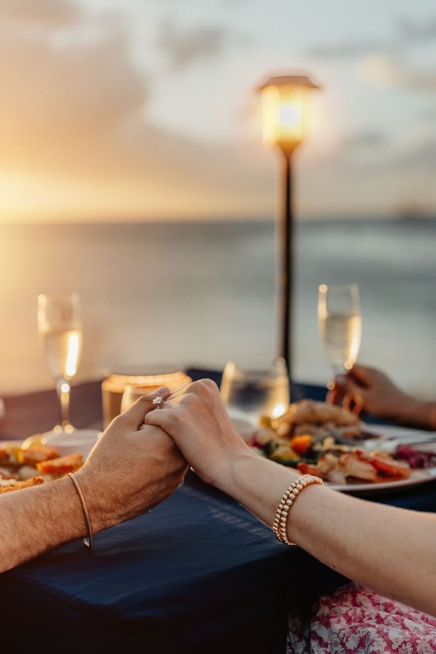 Two people hold hands over a dinner table at sunset with food and wine glasses.