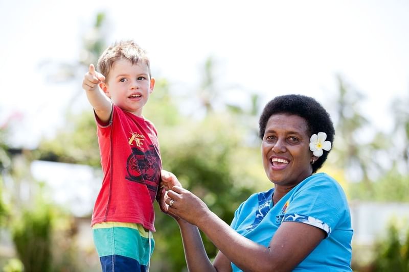 Smiling boy pointing by a woman with a flower under a bright sky near greenery at The Naviti Resort - Fiji