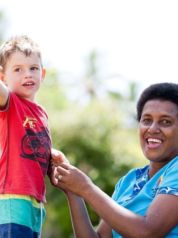 Smiling boy pointing by a woman with a flower under a bright sky near greenery at The Naviti Resort - Fiji