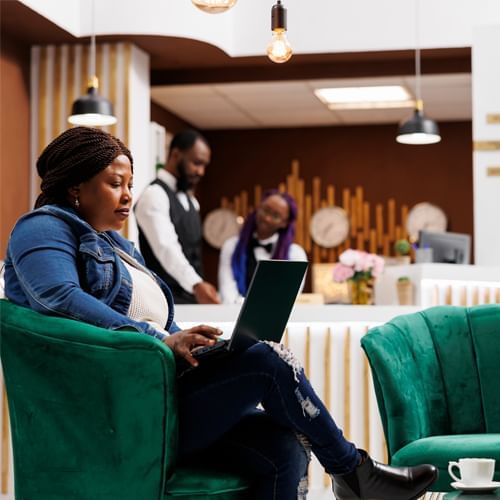 Woman using a notebook on a green lounge chair beside a staffed reception counter in the hotel lobby at Rosen Inn Hotels