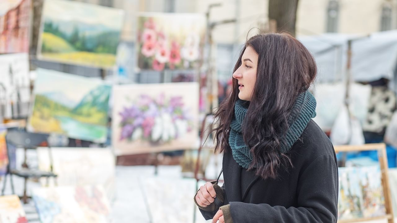 Woman in teal scarf admiring colorful paintings at an outdoor art market.