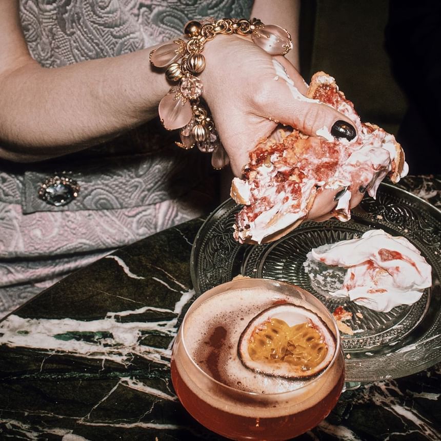 Hand with a bracelet holding a messy dessert beside a cocktail and dessert plate on a marble table at Hotel Motto Vienna