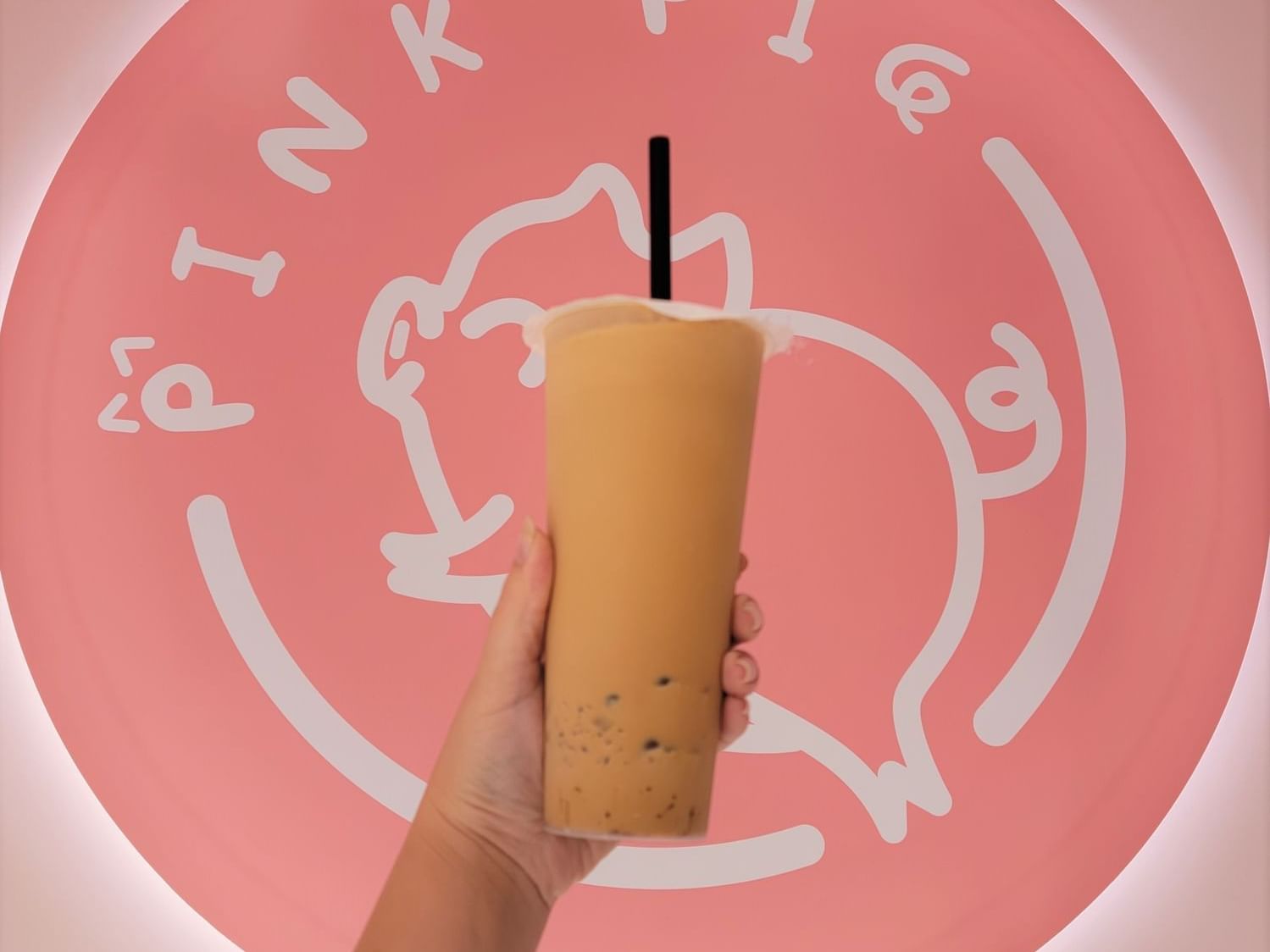 A hand holding a beverage cup in front of Pink Pig Boba & Pizza logo at Grand Legacy at The Park Anaheim.