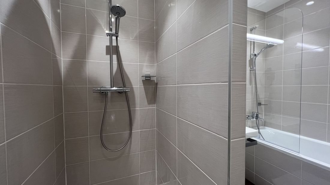 Salle de bain moderne avec douche, carrelage gris, et baignoire visible à travers une porte en verre.