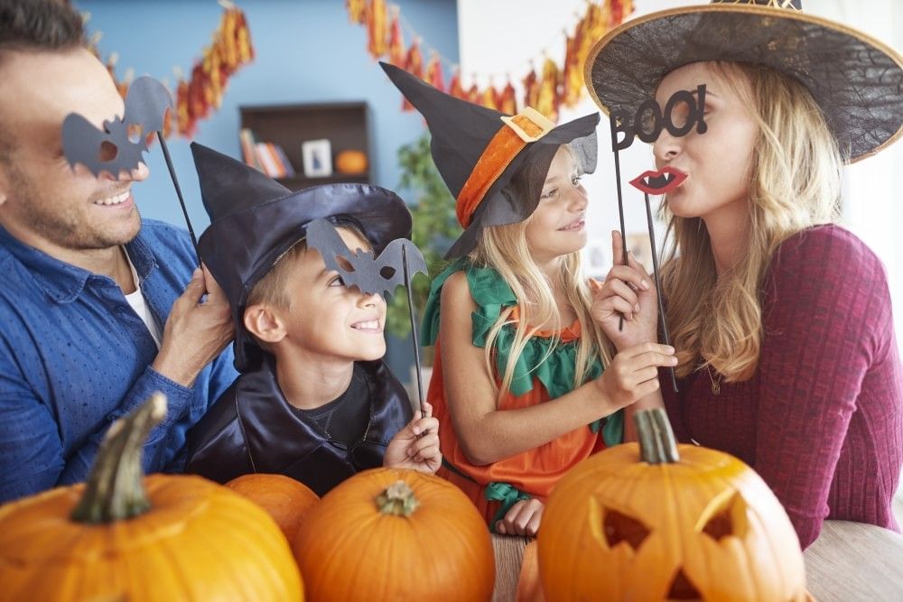 A mom, dad, son, and daughter play with playful masks at a table with carved pumpkins for Halloween.