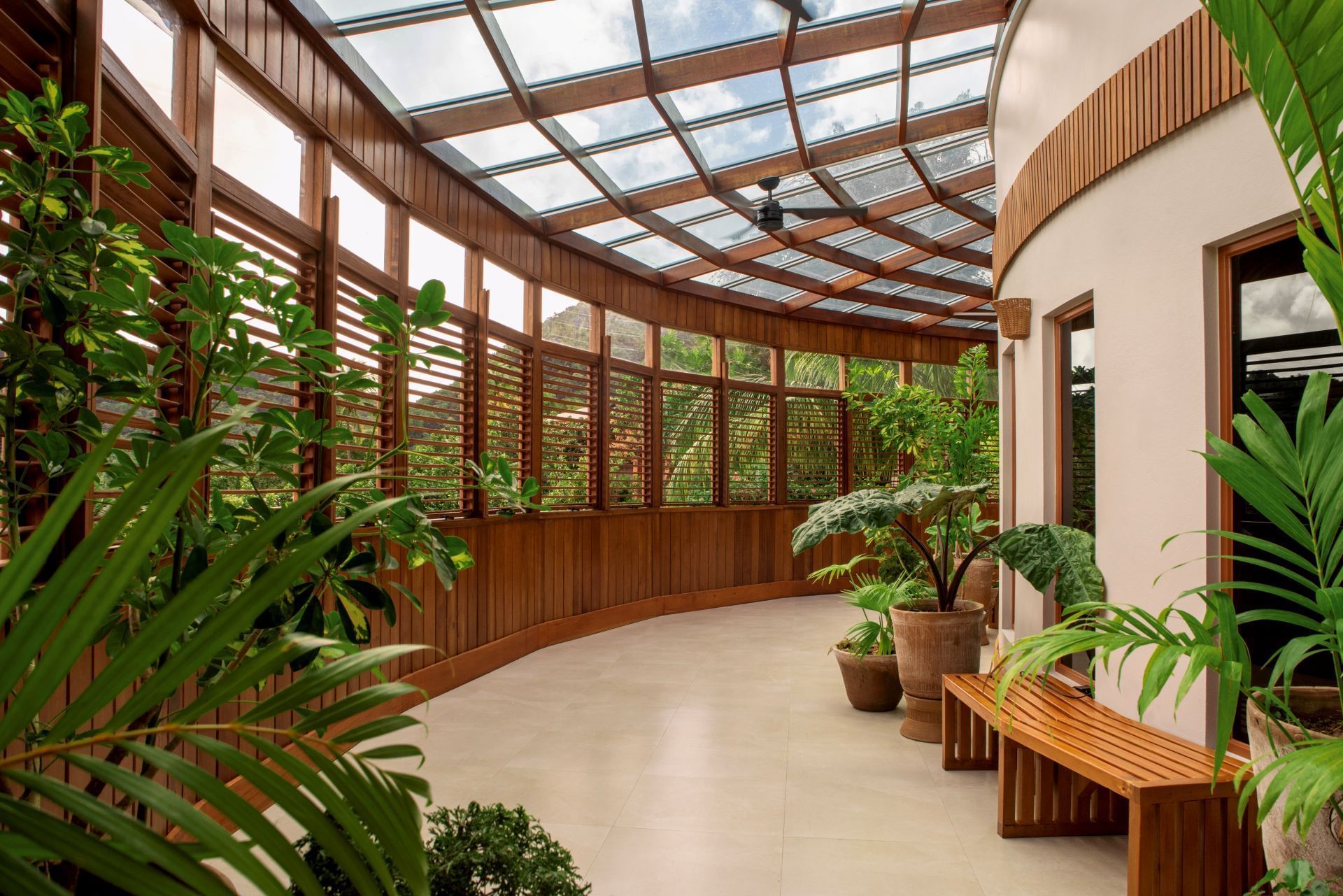 A sunny indoor garden with wooden walls, glass roof, and a wooden bench with various potted plants around.