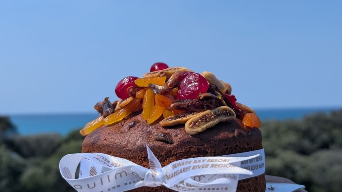 Festive In-Villa Indulgence features a fruit-topped cake on a plate held against a scenic ocean backdrop.