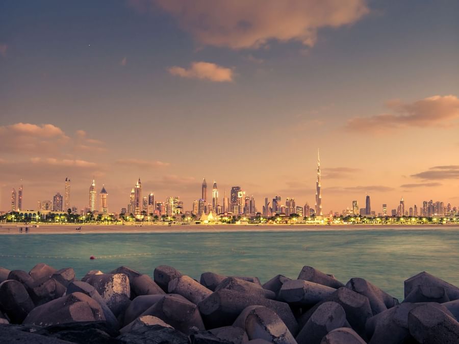 Sunset view of Dubai City skyline across the sea with large rocks near DAMAC Hotels & Resorts