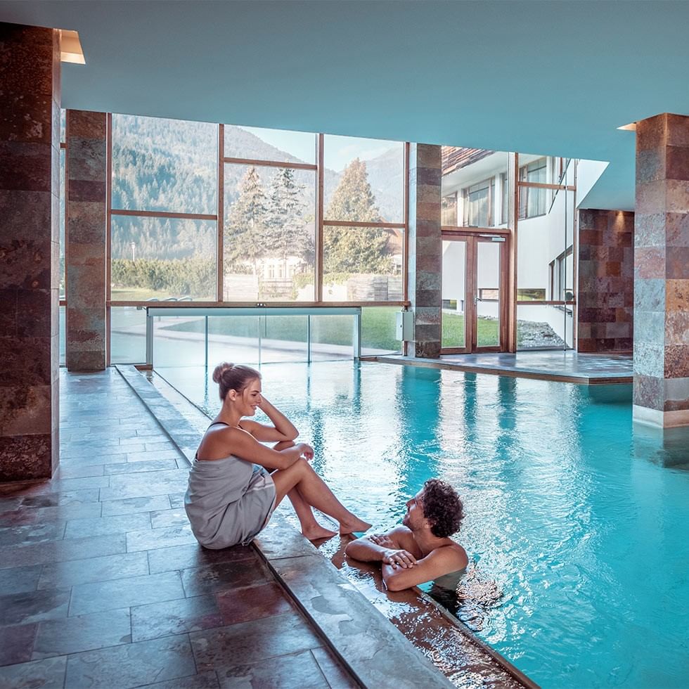 Frau und Mann sitzen in einer modernen Schwimmhalle mit Glaswänden und Bergblick.