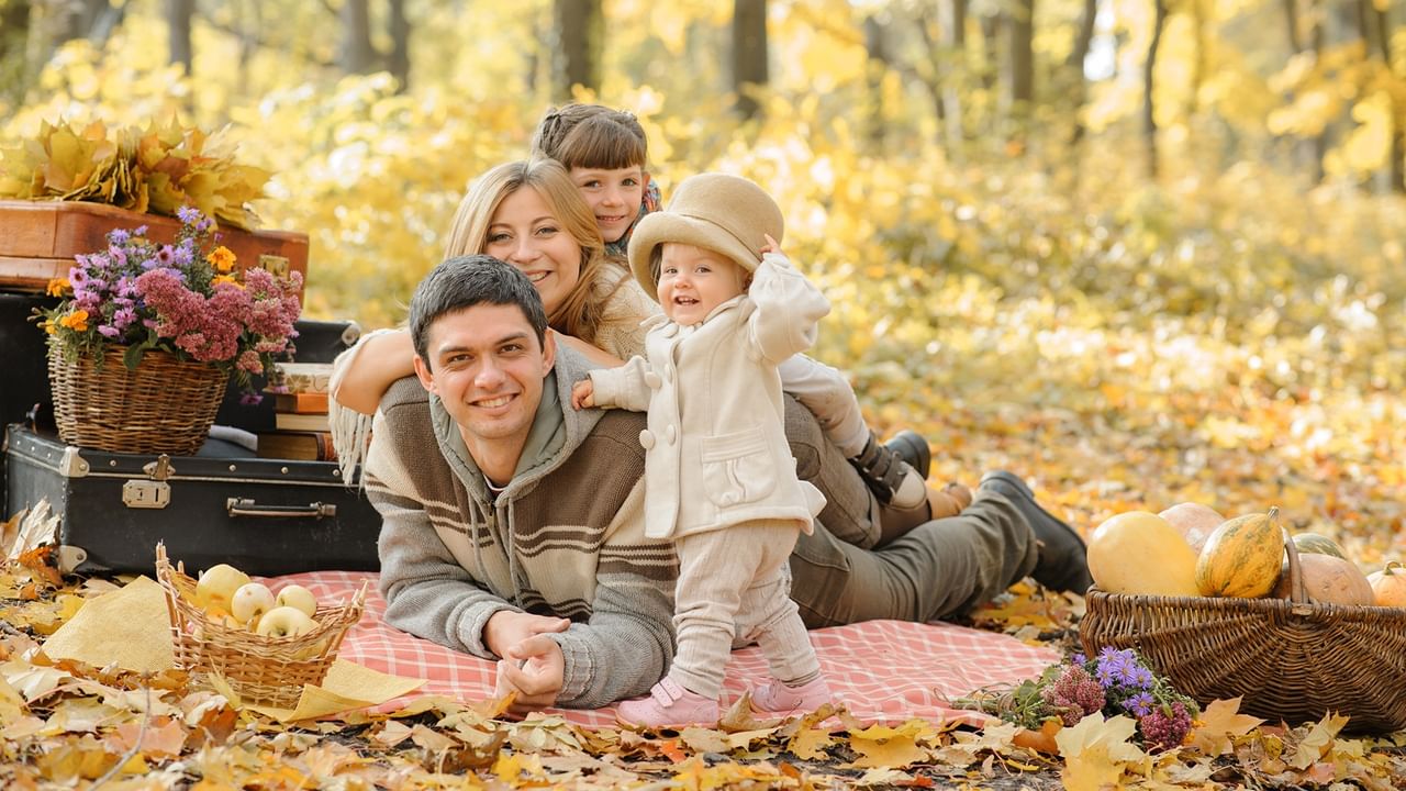 Happy family enjoying a picnic on a blanket surrounded by golden autumn leaves and colorful flowers.