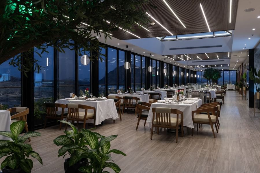 Set tables by glass walls under linear lights surrounding a tree in Karkan Restaurant at Warwick Al Taif Hotel