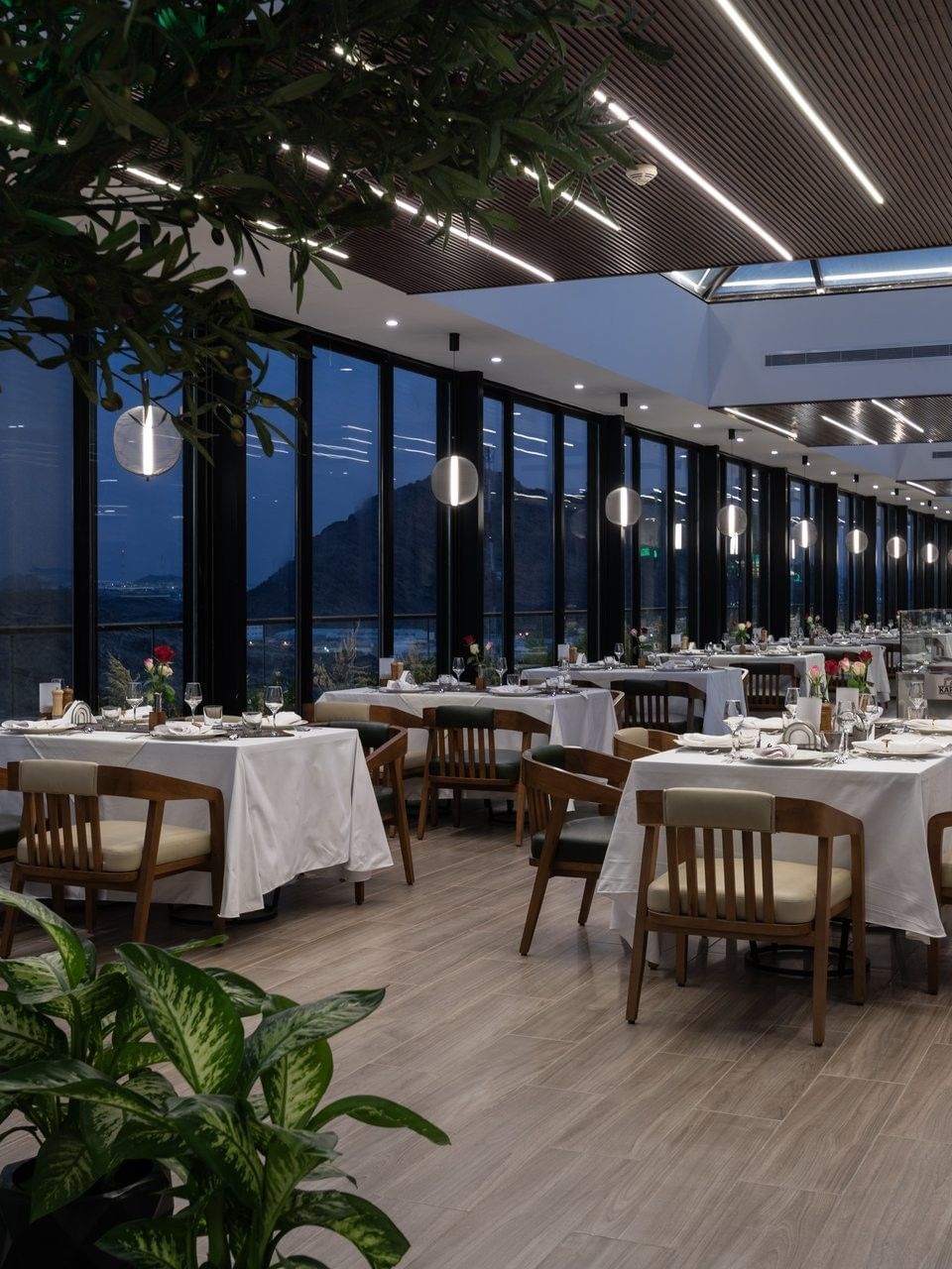 Set tables by glass walls under linear lights surrounding a tree in Karkan Restaurant at Warwick Al Taif Hotel