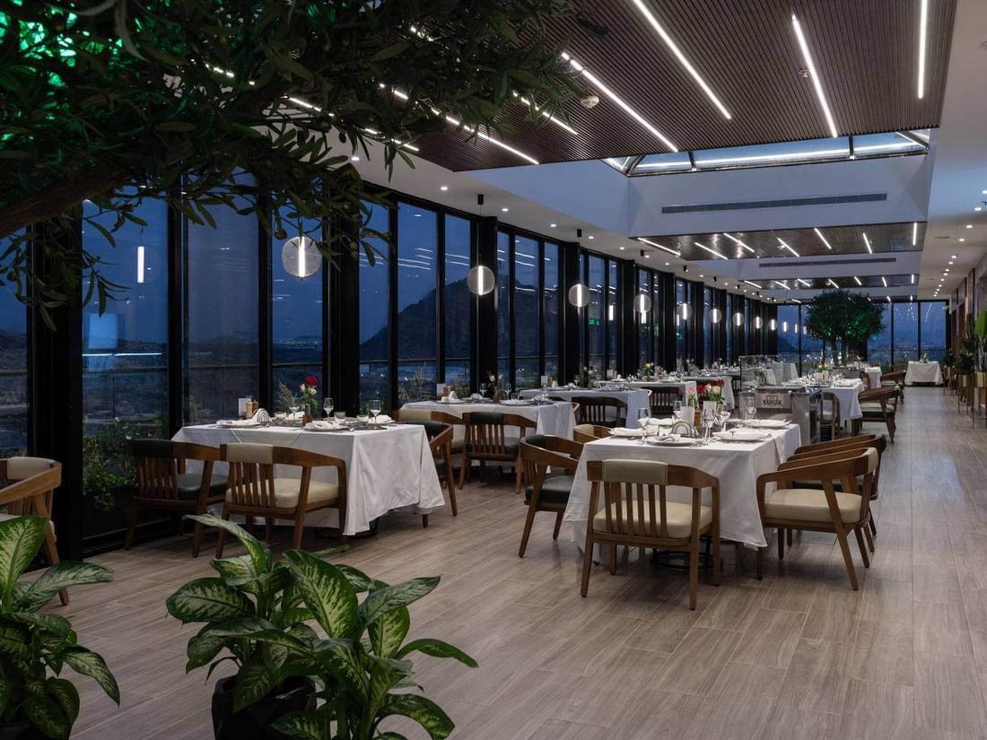 Set tables by glass walls under linear lights surrounding a tree in Karkan Restaurant at Warwick Al Taif Hotel