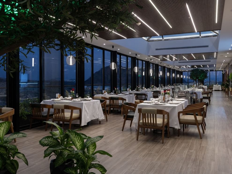 Set tables by glass walls under linear lights surrounding a tree in Karkan Restaurant at Warwick Al Taif Hotel