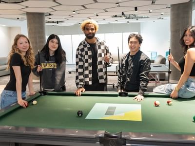 Students smiling in front of pool table in large room with floor to ceiling windows.