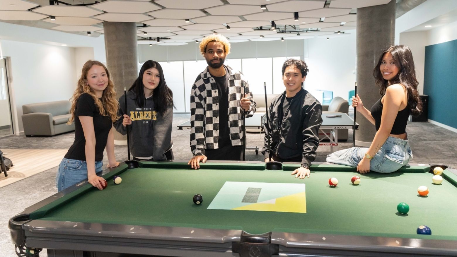 Students smiling in front of pool table in large room with floor to ceiling windows.