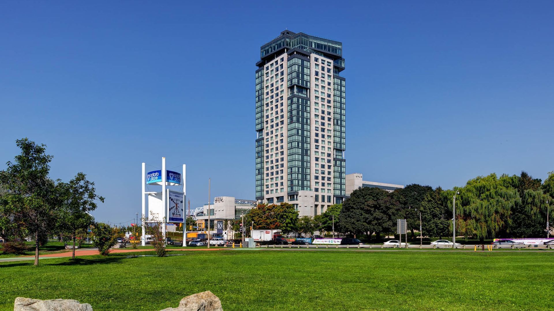 Modern high-rise glass building of Hotel X Toronto rising behind lush green park lawn and trees under clear blue sky