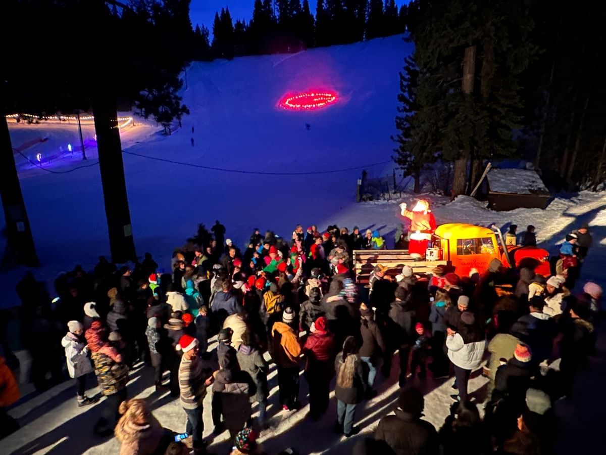 Letter 'G' made with torches burn on ski hill and crowd gathers around Santa on Sno Cat