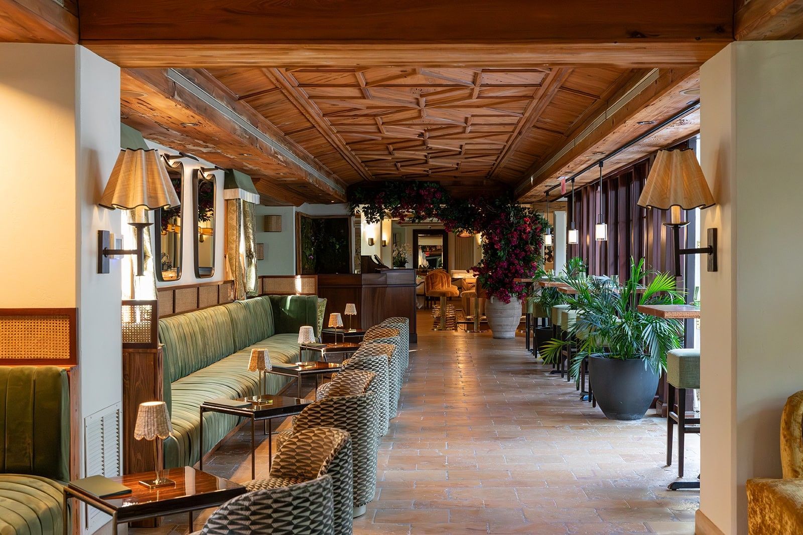 Elegant lounge with wooden ceiling, green benches, patterned chairs, and potted plants along a tiled corridor.