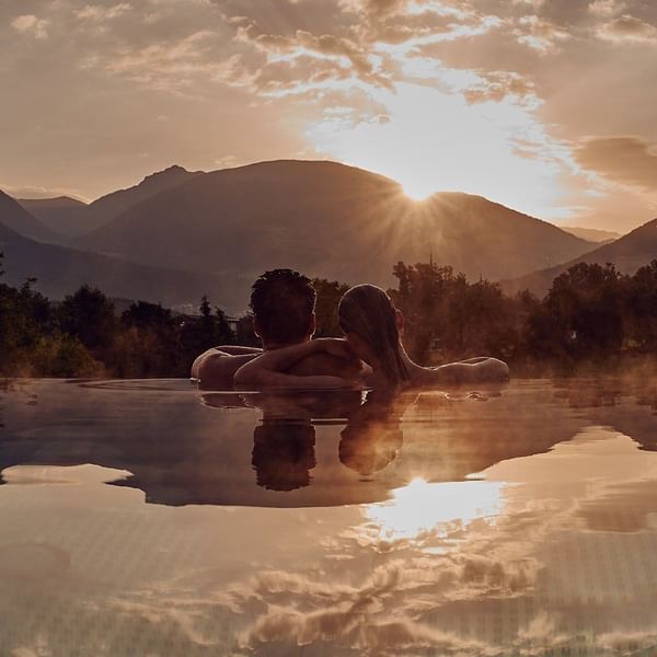 Zwei Menschen genießen einen Sonnenuntergang in einem Pool vor einem Bergpanorama.