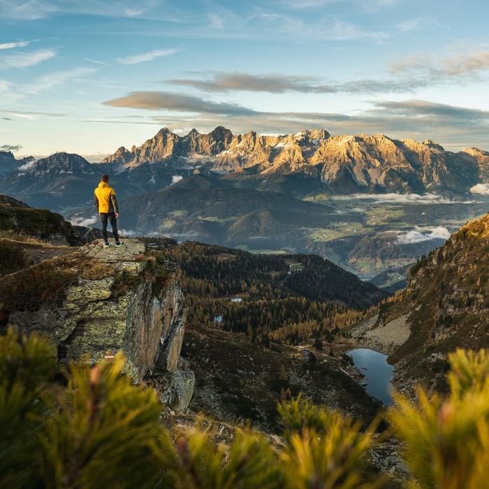Mann auf Felsvorsprung mit Blick auf Berglandschaft und Seen bei Sonnenuntergang.