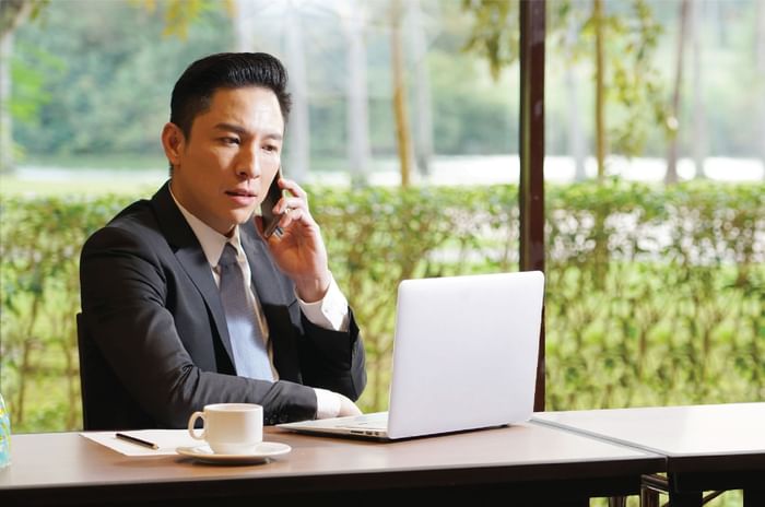 Businessman on a call at a cafe table with a laptop and greenery outside at The Saujana Hotel Kuala Lumpur